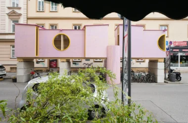 ZuHaus by Clemens Hoyer, pink timber structure on stilts above a Munich street, partially obscured by roadside greenery