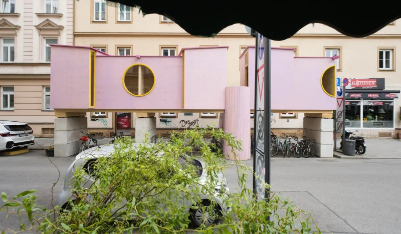 ZuHaus by Clemens Hoyer, pink timber structure on stilts above a Munich street, partially obscured by roadside greenery