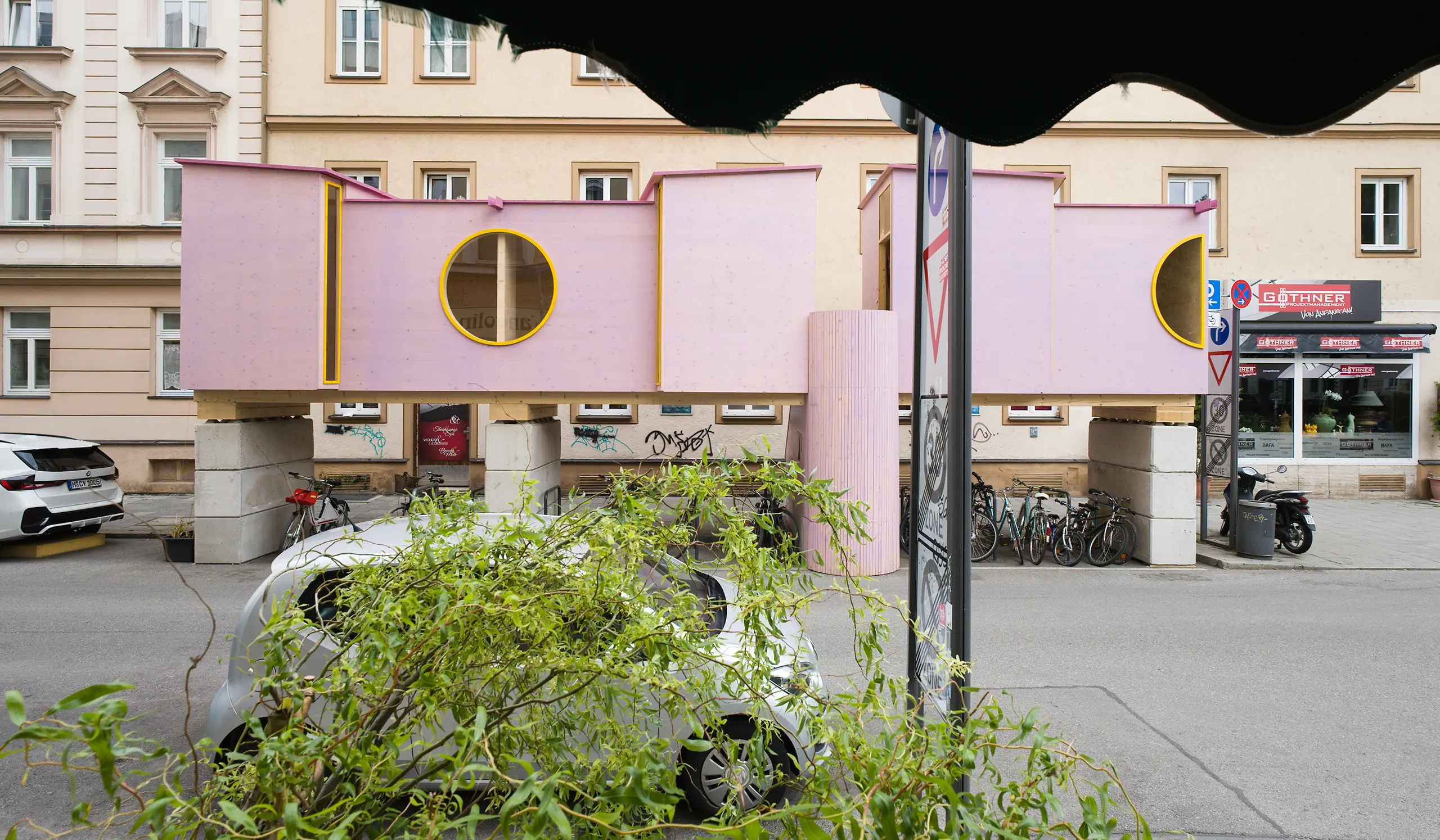 ZuHaus by Clemens Hoyer, pink timber structure on stilts above a Munich street, partially obscured by roadside greenery