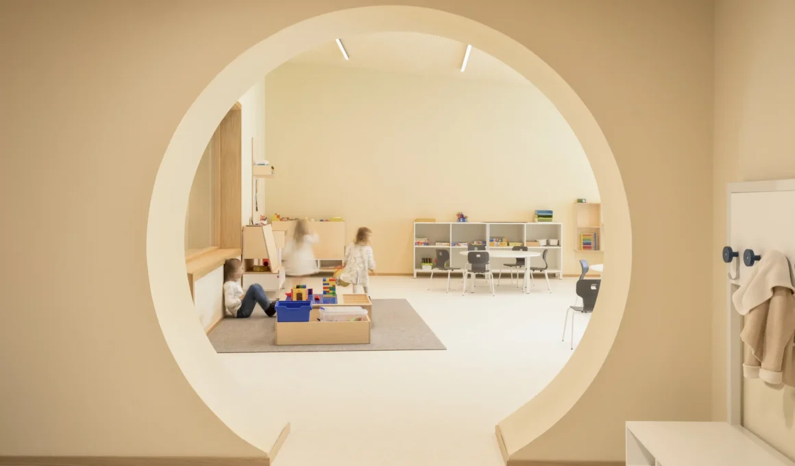 MD41 Longare Children's Center circular doorway framing a play room with wood shelving and round tables. Photography: Alberto Sinigaglia