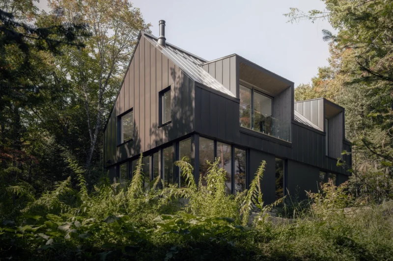 Chalet Moraine by Appareil Architecture: dark gabled volume with recessed dormers and a glazed ground floor in the forest. Photo: Félix Michaud