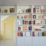 Casa POP by gon architects, opal-green lacquered bookcase with 27 shelves, TV wall, sliding mirror door to yellow bedroom