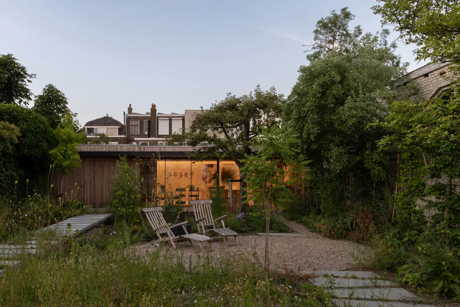 Timber-clad painter's studio volume glowing at dusk, seen across the garden, DP6 architectuurstudio, Delft
