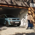 New Dacia Spring in Agave blue charging from a wallbox in a wooden garage, front three-quarter view