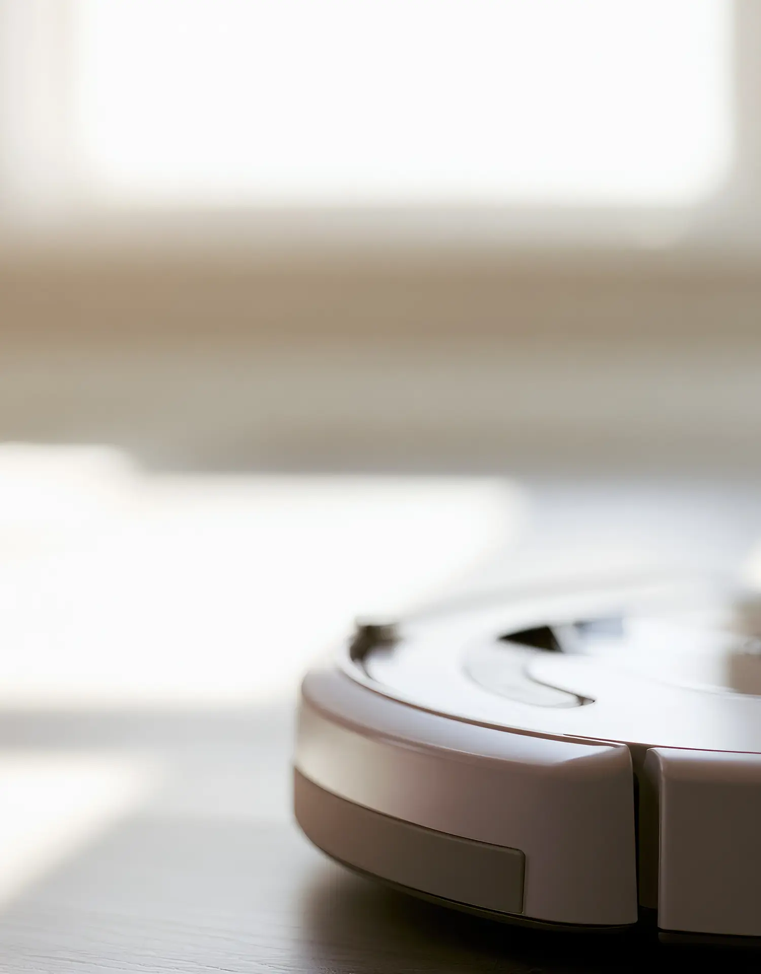 Close-up of the edge of a white robot vacuum on a dark wood floor, with a bright window blurred in the background