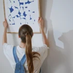 Woman holding a framed blue abstract wall art print against a white wall next to two smaller framed works in thin wood frames