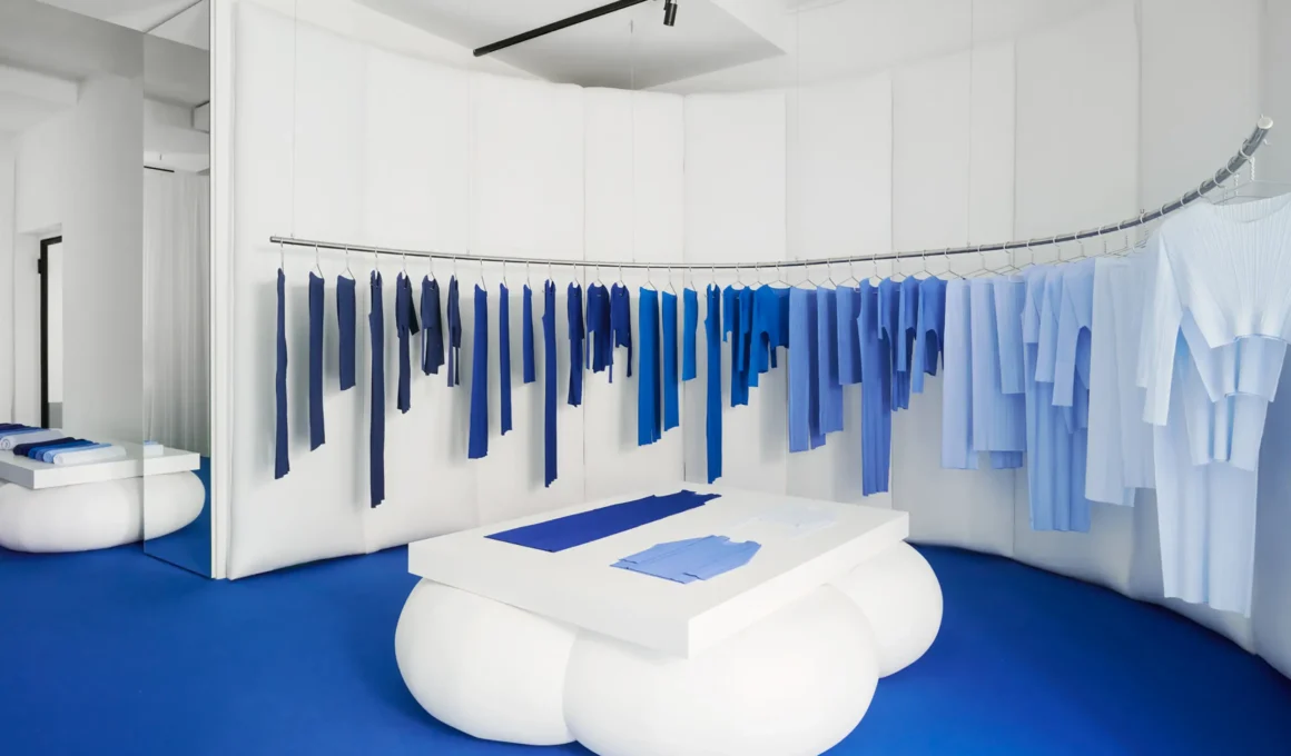 Curved white pleated wall inside the Pleats Please Issey Miyake pop-up at 10 Corso Como, Milan, with Azure Colors garments on a bowed rail over a blue floor