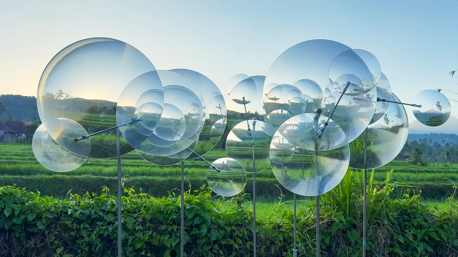 Eccentric Lenses by Vincent Leroy seen head-on at dusk, clustered Fresnel lens discs on vertical poles set against Munduk rice fields, Bali