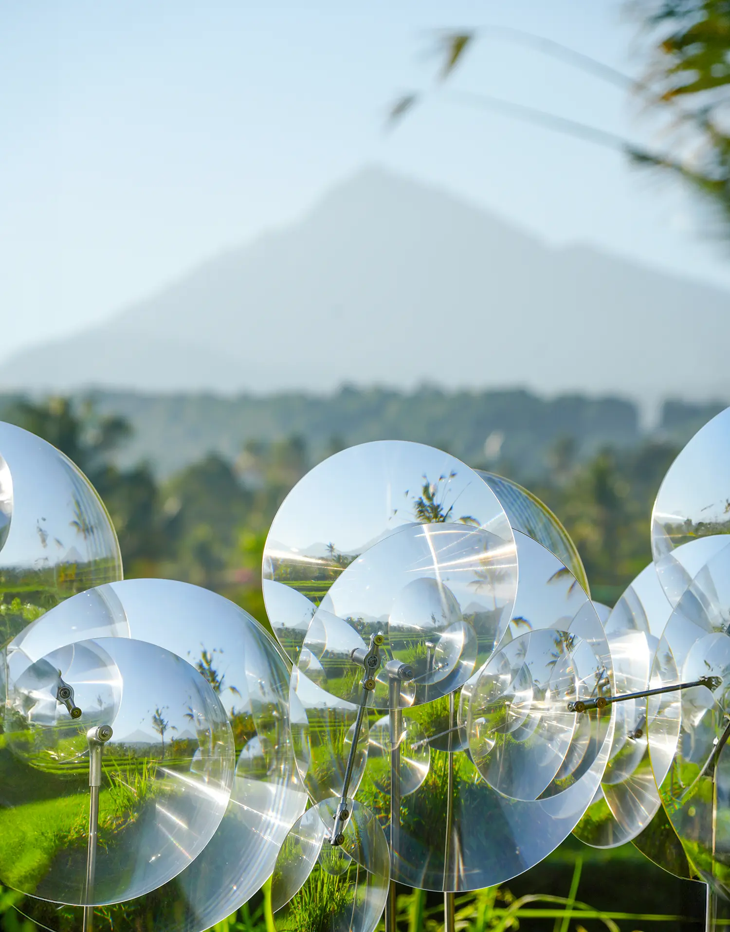 Eccentric Lenses by Vincent Leroy, Fresnel lens discs on poles with a volcanic peak and rice terraces behind, Munduk, Bali