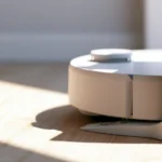 White robot vacuum parked in its docking station on a light wood floor, raking sunlight casting a sharp shadow across the boards